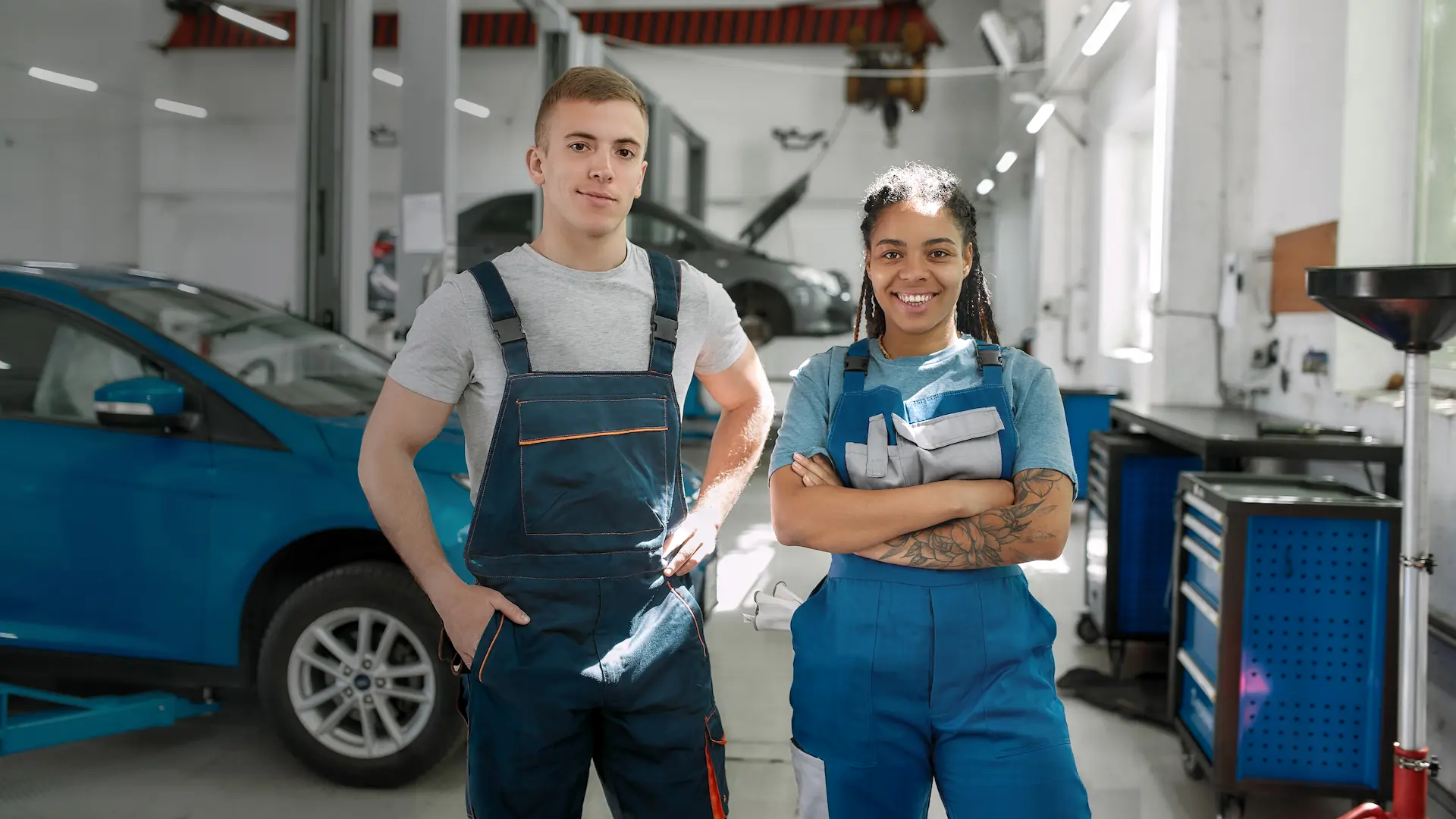 Two young mechanics in overalls inside a well-lit garage.